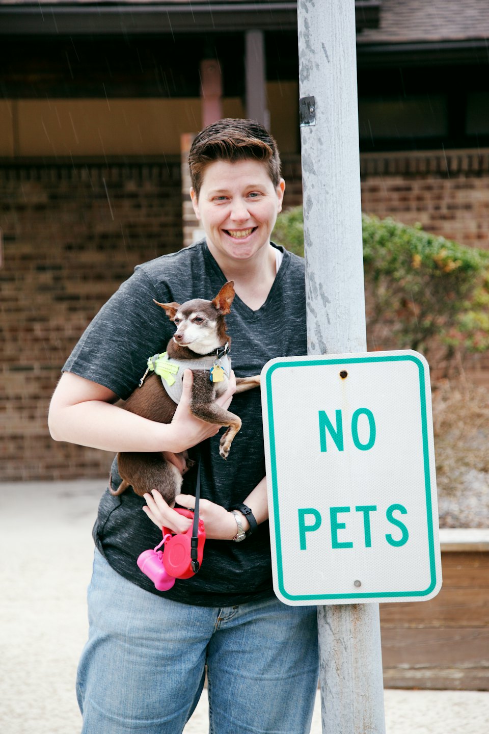 Person smiling with small dog next to 'NO PETS' sign.